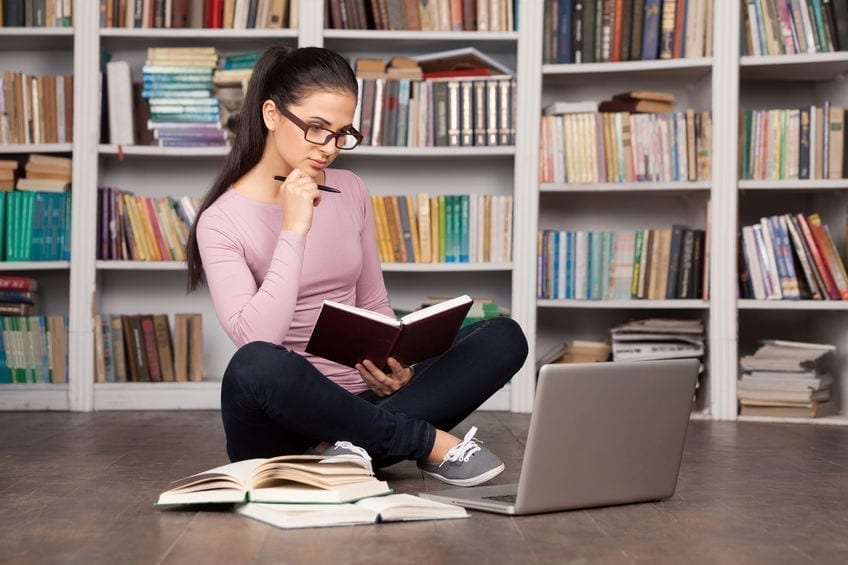 Preparing to the exams. Thoughtful young woman holding a book and looking at the laptop while sitting on the floor at the library